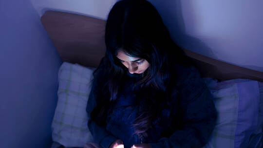 Sad and tired teenager girl using a smartphone lying in bed late at night, playing games, watching videos, and typing. Children'