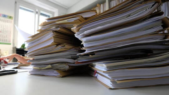 Verwaltungsmitarbeiterin verschwindet fast hinter einem Aktenberg. Das Bild kann symbolisch für den täglichen Papierkrieg mit un KI-Anwendungen in der Verwaltung