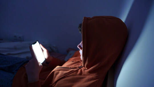 Sad and Tired Teenager Lying in Bed at Night Using Smartphone. Teenager using a smartphone lying in bed late at night, playing