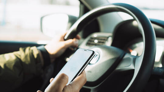 Handling your phone while driving. Close-up of a hand holding a phone and steering wheel of a car while driving.