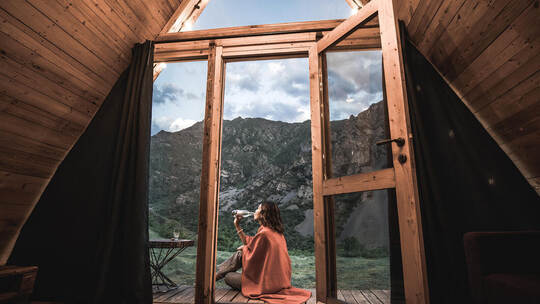 Alone woman drinking water on the porch of a small wooden house in the mountains. The concept of glamping and idyllic holidays