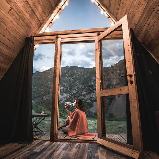 Alone woman drinking water on the porch of a small wooden house in the mountains. The concept of glamping and idyllic holidays