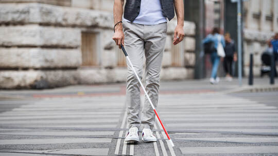Midsection of young blind man with white cane walking across the street in city.