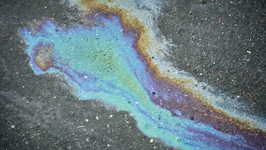A vibrant oil spill spreads across the dark asphalt, revealing shades of blue, purple, and green. The unique patterns draw atten