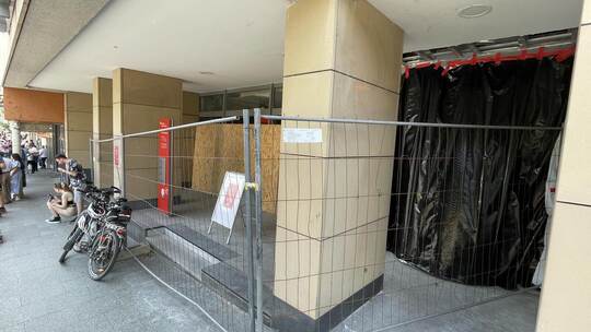 Der Eingang und die Fenster sind mit Holz verkleidet, das Gebäude zusätzlich mit einem Bauzaun gesichert. Außen hängen Hinweissc
