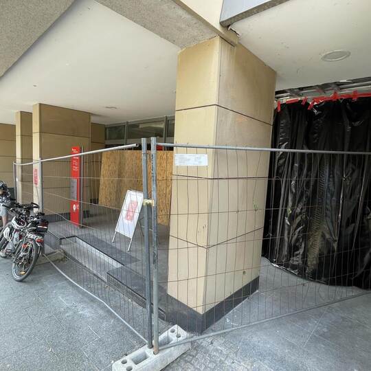 Der Eingang und die Fenster sind mit Holz verkleidet, das Gebäude zusätzlich mit einem Bauzaun gesichert. Außen hängen Hinweissc