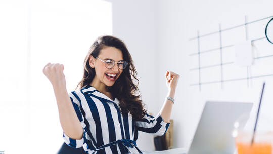 Happy young woman sitting at table with hands up in winner gesture