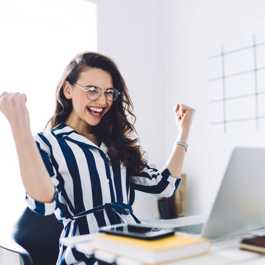 Happy young woman sitting at table with hands up in winner gesture