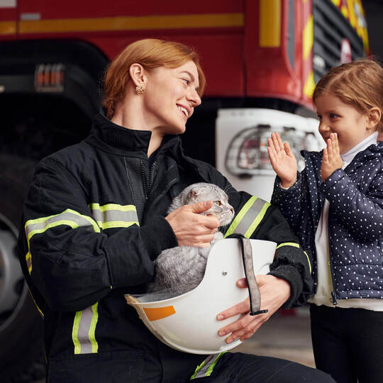 Cat is sitting in the helmet, kid is happy, clapping her hands. Firefighter woman in uniform is with a little girl