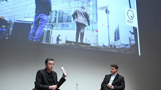 Robert Eikmeyer (links) und Tobias von Laubenthal im Gespräch. Über ihnen zeigt ein Videoausschnitt das „Walter Lübcke Memorial“