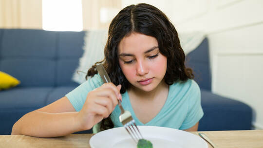 Sad teenage girl staring at a lone broccoli on a plate, struggling with anorexia or bulimia and maintaining a highly limited die