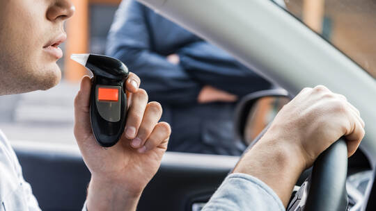 cropped view of driver blowing into breathalyzer, and policeman standing near car on blurred background.