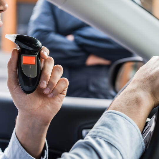 cropped view of driver blowing into breathalyzer, and policeman standing near car on blurred background.