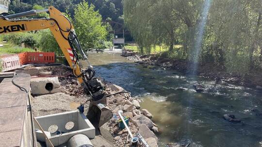 Der Bagger steht im Flussbett des Reichenbachs. Auf beiden Seiten sind die Löcher des Abwasserkanals zu sehen. Rechts unten im B