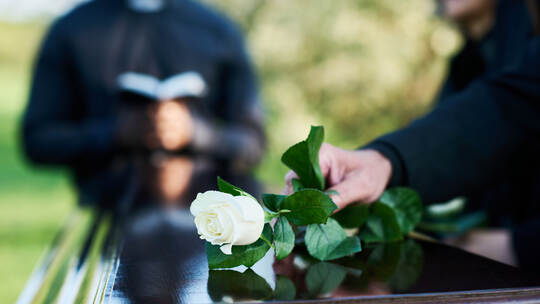 Fresh white rose being put by woman in mourning attire on closed coffin lid