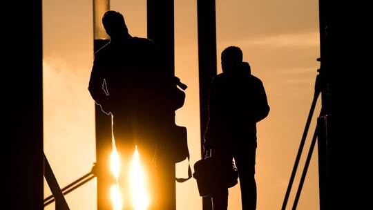 Zwei Männer gehen mit Aktentasche bei Sonnenaufgang über Brücke Zwei Männer gehen mit Aktentasche bei Sonnenaufgang über Brücke