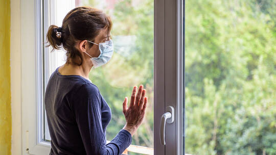 A woman wearing a medical face mask is staring into space through a window.