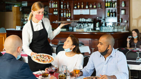Waitress in protective mask bringing ordered pizza to guests