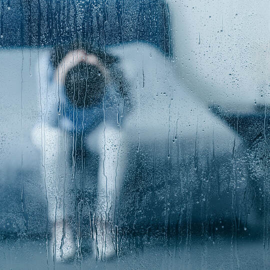 depressed woman sitting on bed and holding head in hands through window with raindrops