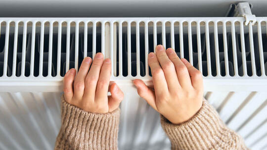 Child wearing brown woolly jacket warming cold hand in front of heating radiator in winter time. Electric or gas heater at home.